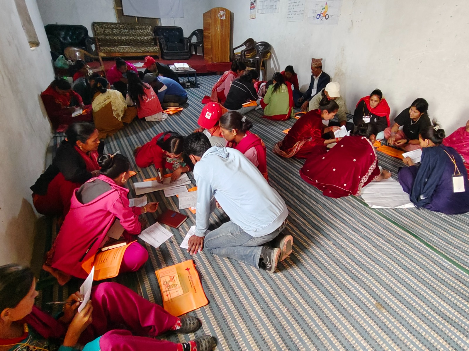 A group of volunteers and community members sit on the floor of a room, filling out orange Nepal Red Cross Society forms during a Behaviour Change and Impact (BCI) training session.