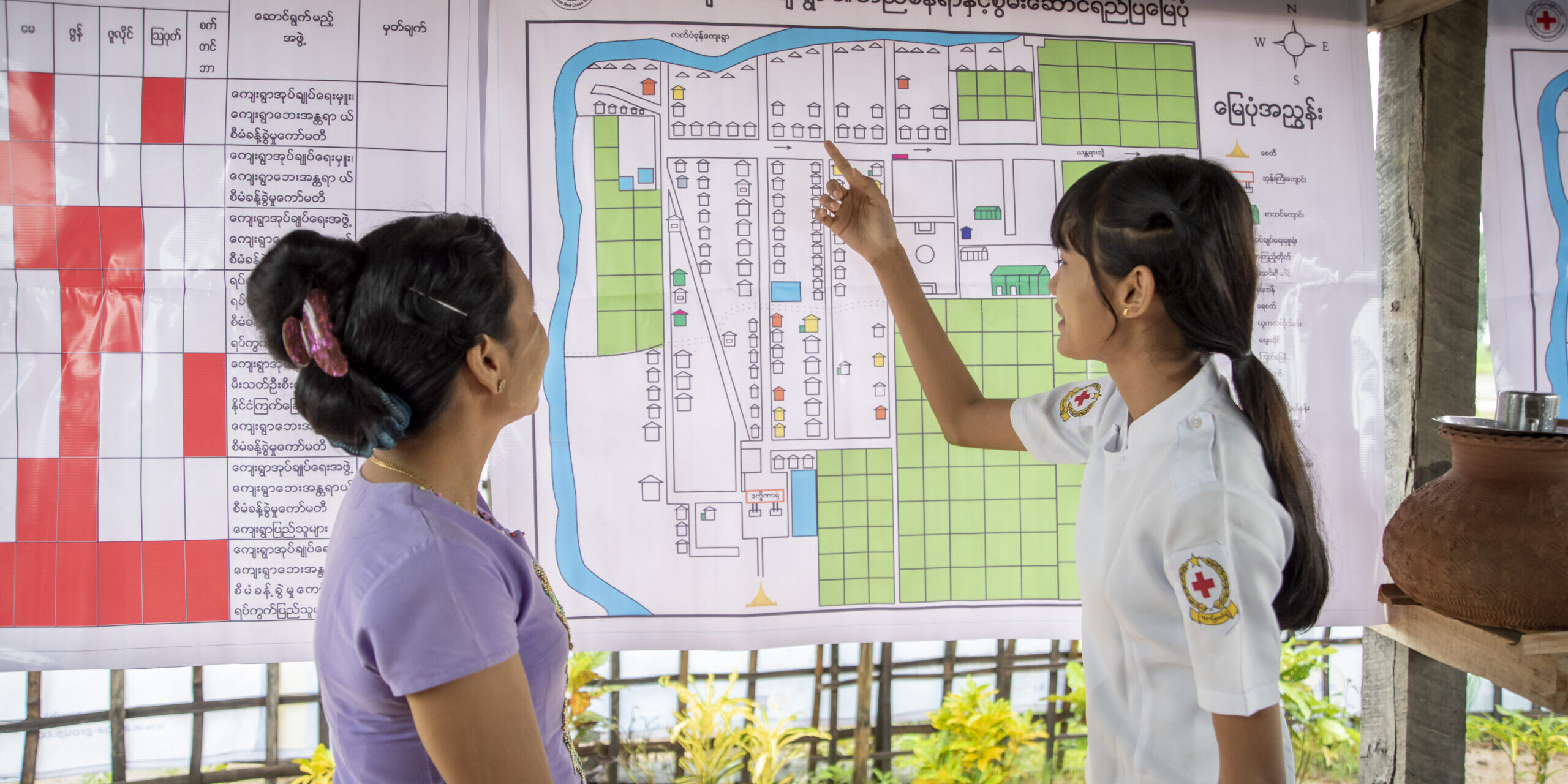 July 9, 2019. Kim Chaung village, Bago, Myanmar. Residents (Ma Sandar Win and Ma Han Thae Oo) of Kim Chaung village, Myanmar discuss a community map hanging in the town center. Developed with help from the Red Cross, the map can be used to mitigate disaster risks. Communities like this one work together to identify their risks—such as flood zones— as well as resources they can utilize during disasters. Maps show homes, high-risk areas, evacuation routes, locations of handicapped residents and the elderly (who may need help evacuating), and safe spaces to seek shelter. This rural community—dotted with rice patties and farmland—experiences regular floods and is at-risk of earthquakes and strong storms.

The American Red Cross works alongside the Myanmar Red Cross to prepare disaster-prone communities for cyclones, floods, tsunamis, earthquakes, and other emergencies. We train and equip families with the tools they need to mitigate natural disaster risks and to be first responders when crises strike. In Myanmar, the American Red Cross teaches basic first aid, light search-and-rescue, and post-disaster epidemic control in 20 communities—in addition to running disaster simulations and forming village committees who mobilize when disasters hit. In 24 schools, we teach students basic first aid, light search-and-rescue, evacuation activities, and distribute emergency equipment—such as solar panels, fire extinguishers, megaphones, early warning speakers, first aid kits, and helmets. In Myanmar, some American Red Cross project sites are urban, while others sit in river deltas only accessible by boat. Note: The country of Myanmar is also known as “Burma.” Photo by Brad Zerivitz/American Red Cross