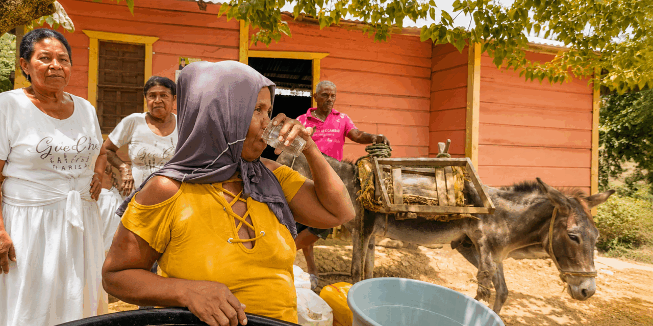 A woman in a yellow top and head covering drinks from a glass of water outside a coral-colored wooden house, with two women, a man, and a donkey carrying a wooden saddle in the background under bright sun.