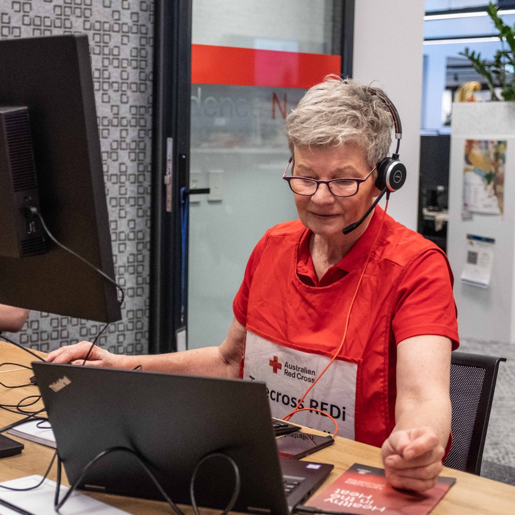 Australian Red Cross volunteer wearing a headset and Redi program vest, seated at a desk with two computer monitors, assisting remotely during a heatwave emergency response