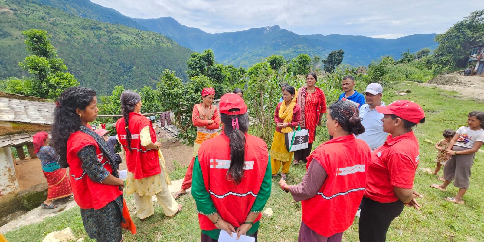 Nepal Red Cross Society volunteers in red vests and caps stand in a circle with community members on a hillside, discussing disaster risk reduction (DRR) against a backdrop of green mountains.