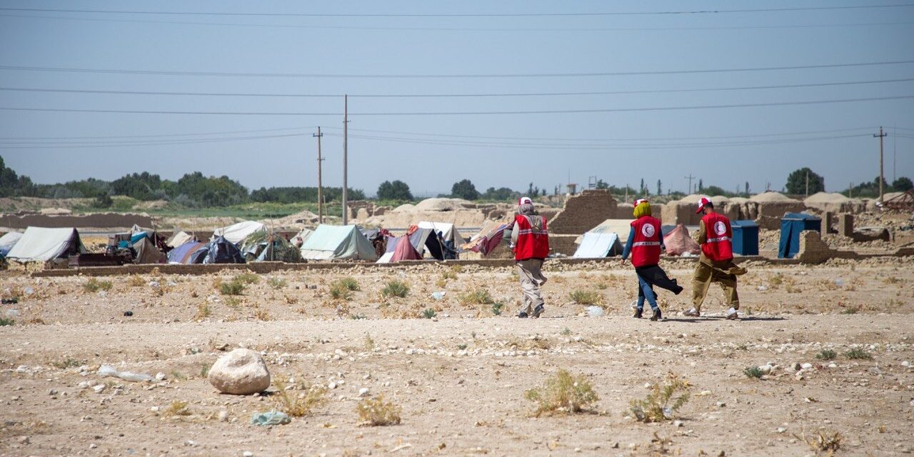 Afghan Red Crescent Society volunteers in red vests walk across a dry, arid landscape towards a temporary displacement camp with rows of tents in Afghanistan.