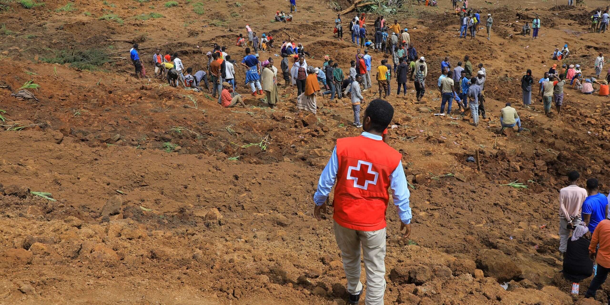 An Ethiopian Red Cross volunteer in an orange vest walks towards a large crowd of people gathered at the site of a landslide in Ethiopia.