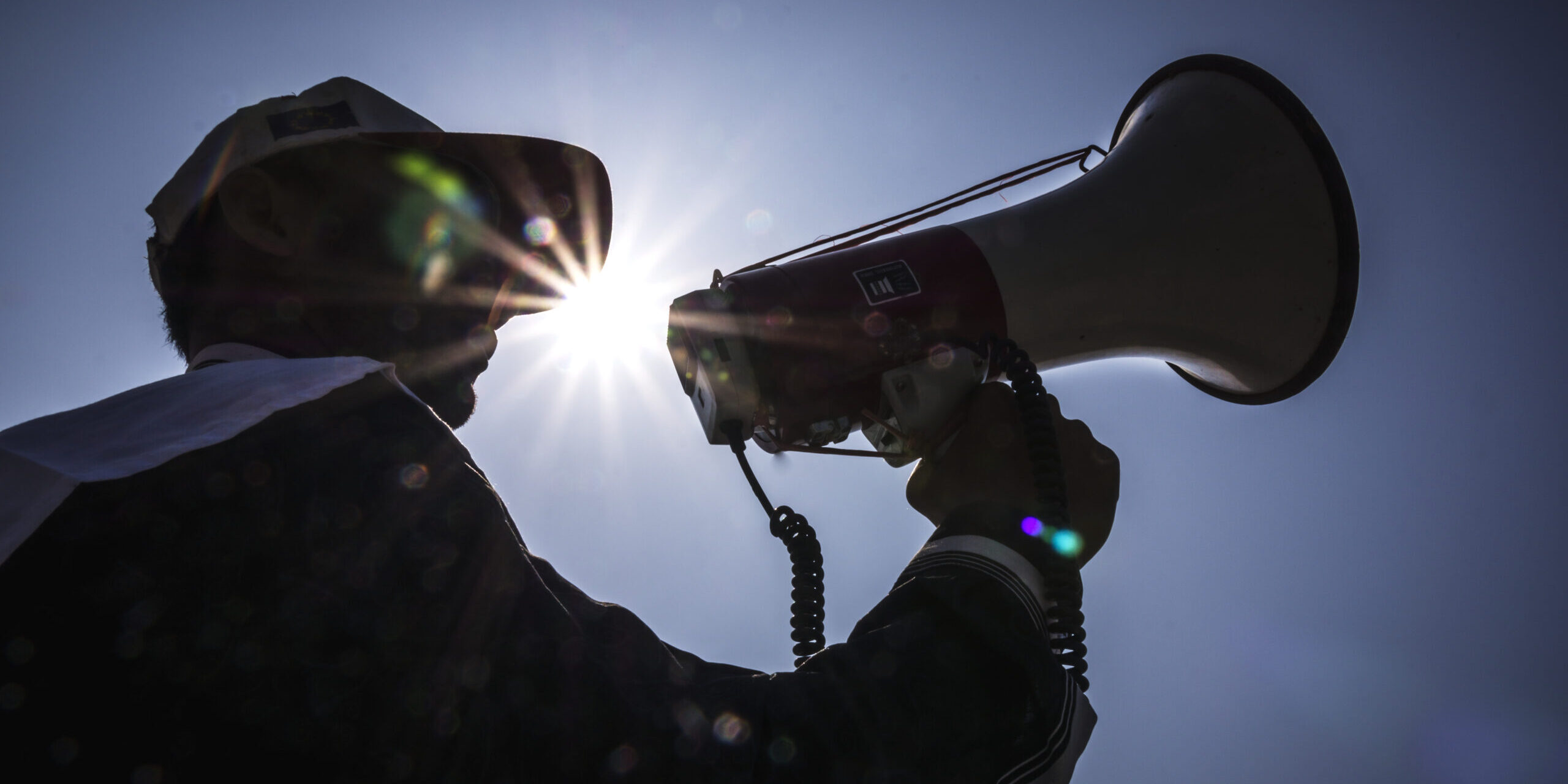 A silhouetted community volunteer uses a megaphone to deliver an early warning message outdoors, with sunlight behind them against a clear sky.